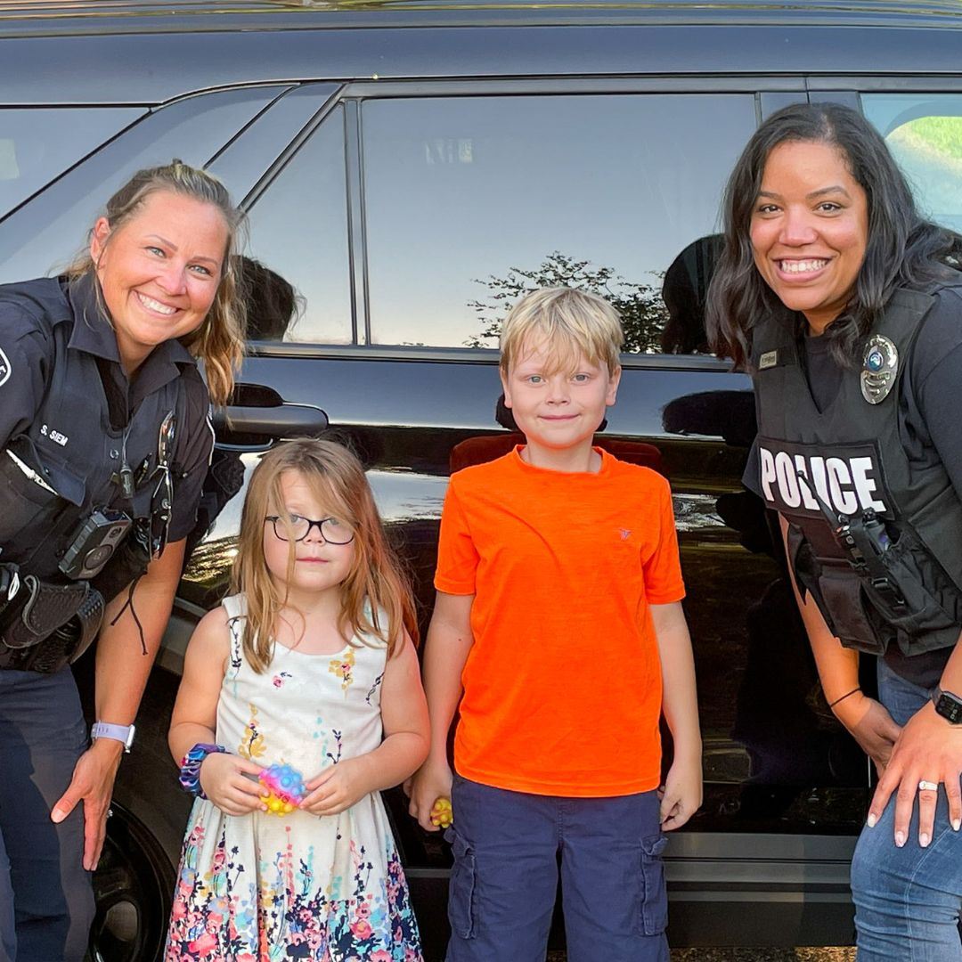 Two police officers and two kids smile in front of a squad car