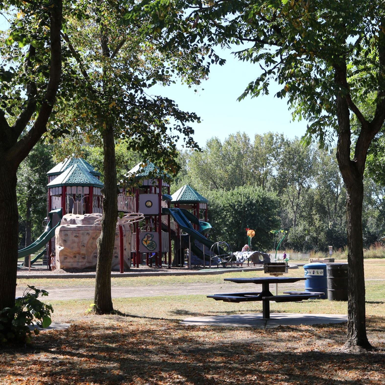 City park with large trees, picnic table, and large playground. 