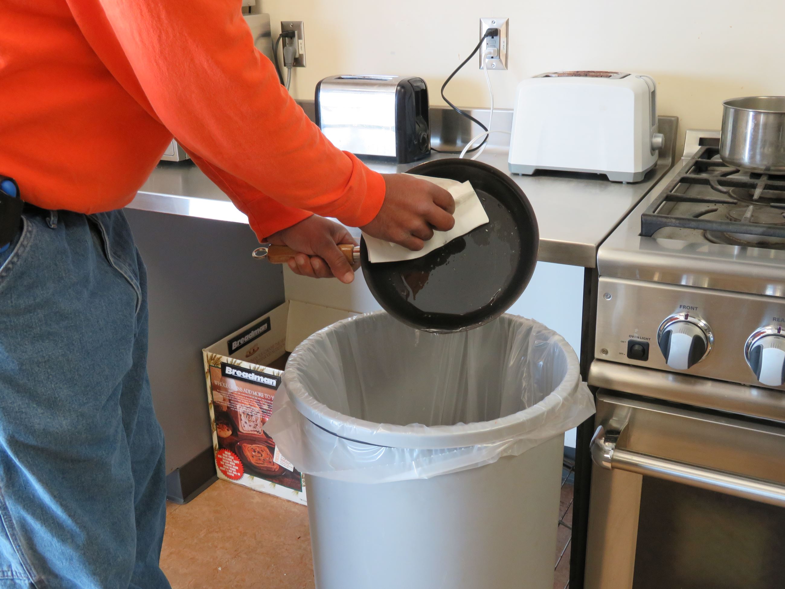 Person scraping grease with a paper towel from a pan into a trash bin.